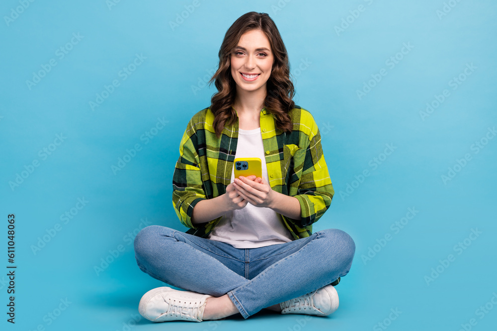 Full body portrait of positive lovely lady sit floor use smart phone isolated on blue color background Full body portrait of positive lovely lady sit floor use smart phone isolated on blue color background