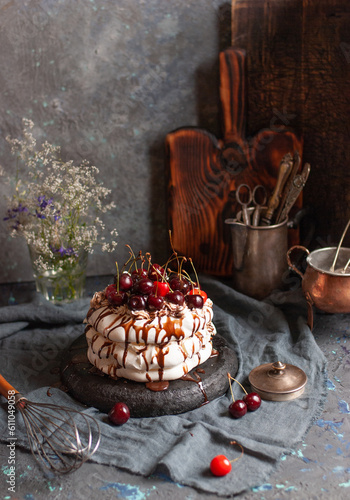 Homemade meringue cake with cherries and chocolate syrup on a dark background surrounded by vintage crockery and flowers