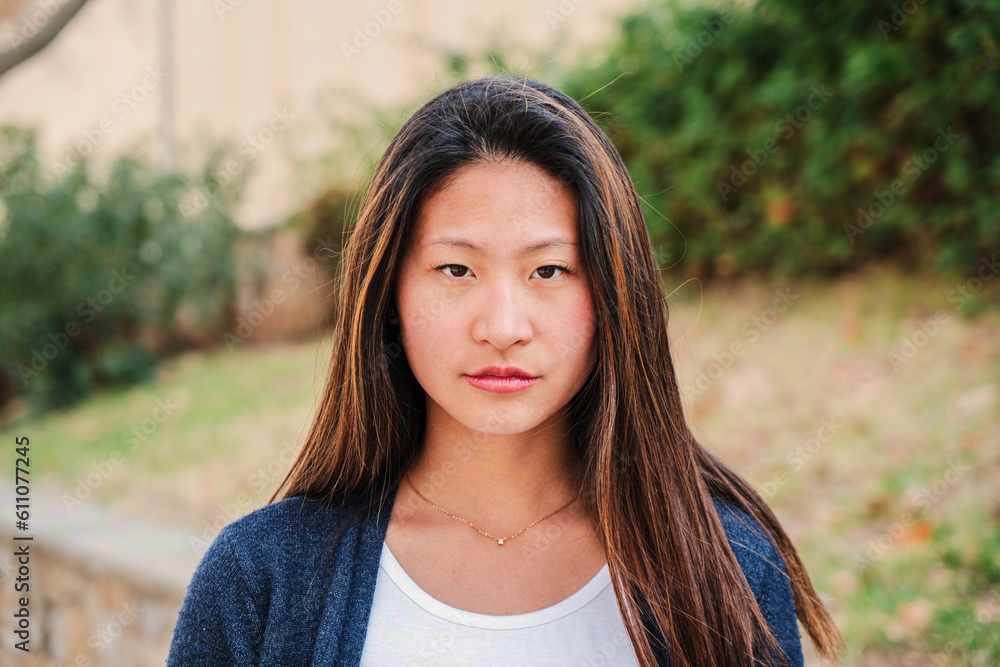 Individual close up portrait of serious young woman standing outdoors. One pensive teen female student looking at camera. Front view of a young chinese lady. Head shot of a proud teenage school girl