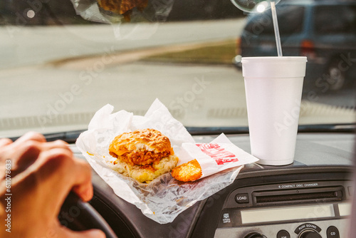 A chicken biscuit and hash browns with a styrofoam cup on the dashboard of a car with a hand on the steering wheel photo