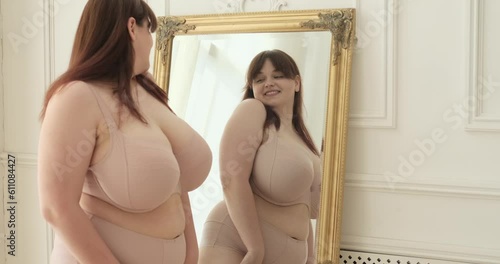 A curvy Caucasian woman standing beside a mirror in her bedroom, engaging in a moment of self-reflection. With a genuine smile and a sparkle in her eyes, she embraces her image.