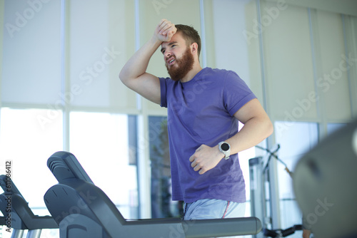 Tired exhausted young man feeling bad, unhealthy, unwell during run on treadmill in the gym holding his head, having headache 