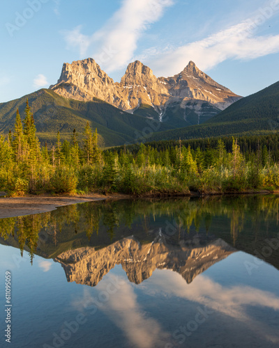 Three Sisters in Canmore seen at golden hour, sunset on blue sky day, afternoon with calm, peaceful reflection in water below famous, tourist, tourism mountains, area summer. 