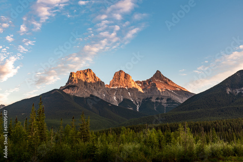Sunset wilderness views in Canmore during summer with Three Sisters in view. Golden glow on mountain peaks. Background, wallpaper, desktop scenic view, tourist, tourism area. Blue sky, clouds