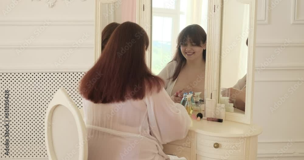 Confident plus size Caucasian woman seated in her bedroom near a vanity table. She gracefully applies cosmetics, touching her face, enhancing her natural beauty. She in good mood and smiling. Confident plus size Caucasian woman seated in her bedroom near a vanity table. She gracefully applies cosmetics, touching her face, enhancing her natural beauty. She in good mood and smiling.