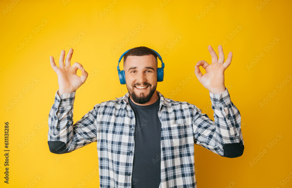 Naklejka premium Happy caucasian guy shows a good gesture, agrees with something, claims that everything is fine, smiles broadly, wears headphones, enjoys isolated on a yellow background