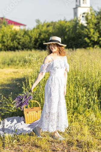 Beautiful young girl in a white dress, straw hat, with picnic and bouquet of purple wild flowers on a meadow. Summertime, golden hour, sunset