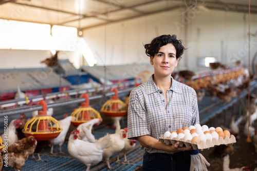 Happy successful hispanic female owner of poultry farm engaged in breeding of laying hens standing in henhouse, holding cardboard tray of fresh organic chicken eggs