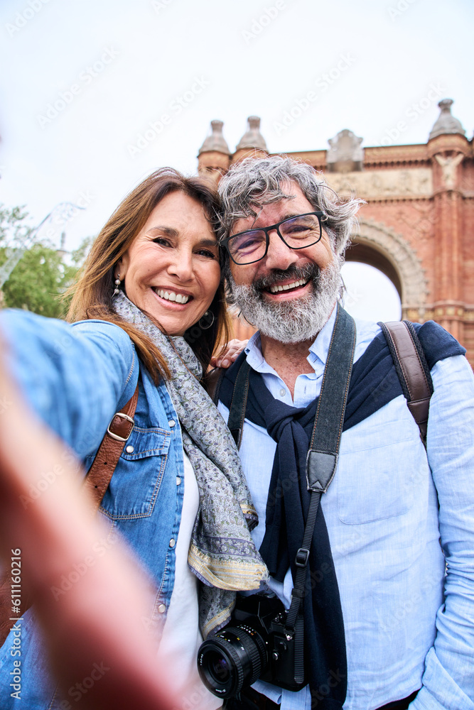 Vertical portrait happy senior tourist couple taking selfie outdoors in historical city. Smiling elderly people traveling together on vacation. Cheerful man and woman posing for photo mobile phone.