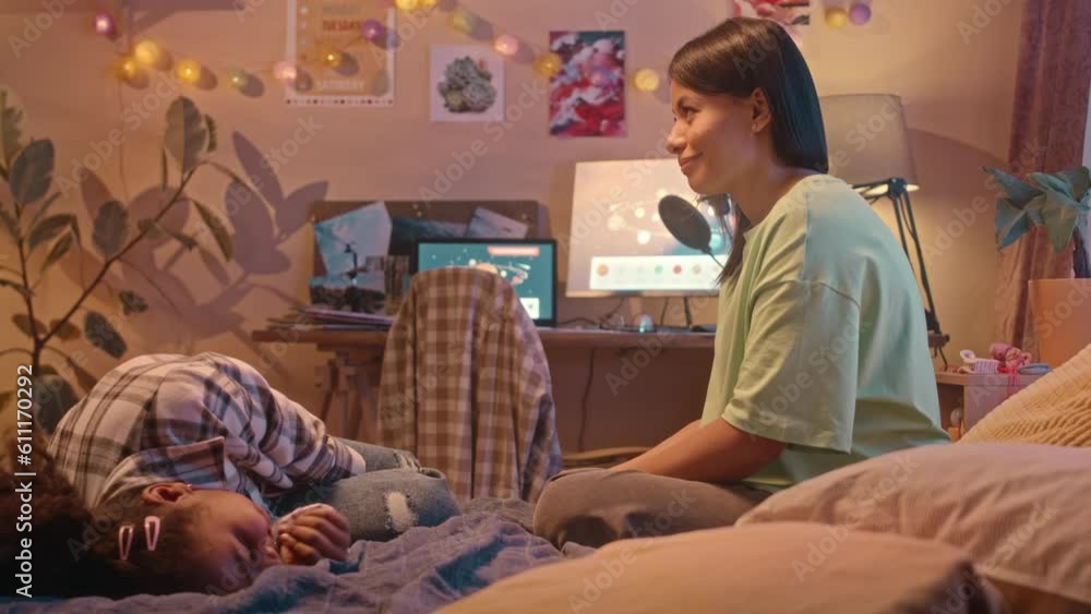 Medium shot of young Biracial woman and her cool preteen daughter sitting on bed in cozy kids bedroom late at night having conversation Medium shot of young Biracial woman and her cool preteen daughter sitting on bed in cozy kids bedroom late at night having conversation