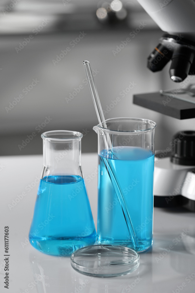 Different laboratory glassware with light blue liquid near microscope on table Different laboratory glassware with light blue liquid near microscope on table