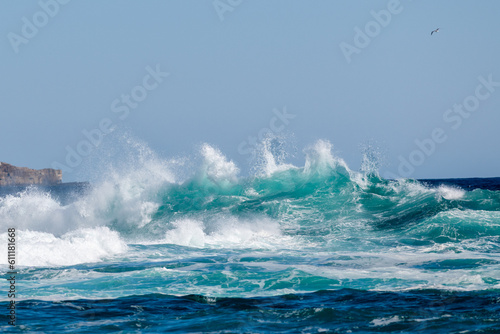 A teal or turquoise colored rip curl of a wave crashing on shore. The dangerous ocean swell has white foam and a water spray. There's a break in the center of the stormy surf with froth on both sides.