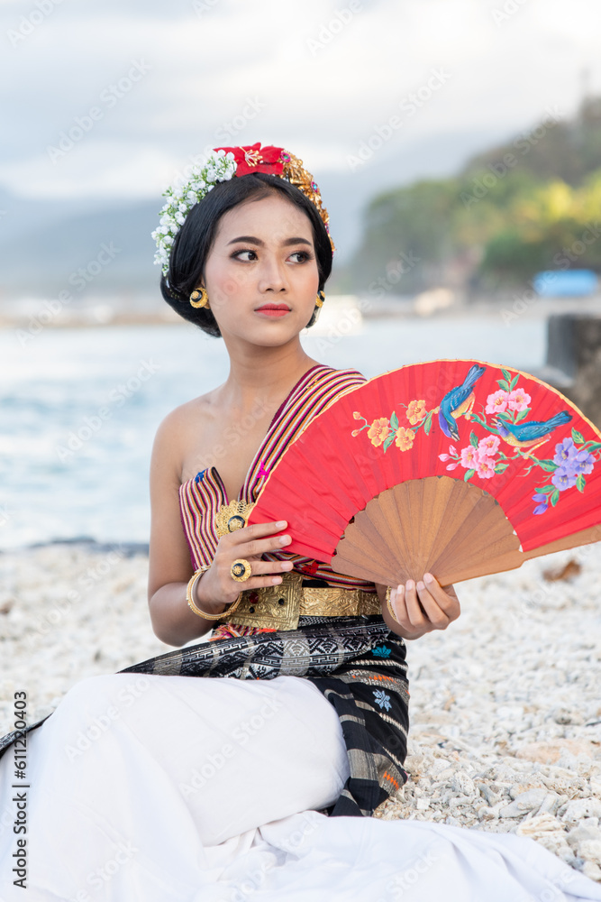 Fototapeta premium Portrait of a cute girl in a beautiful off-the-shoulder costume and a crown in her hair posing by the seashore with red hand fan