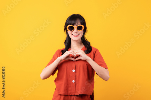 Portrait young beautiful asian woman happy smile dressed in orange clothes and sunglasses showing heart hand gesture isolated on yellow studio background.