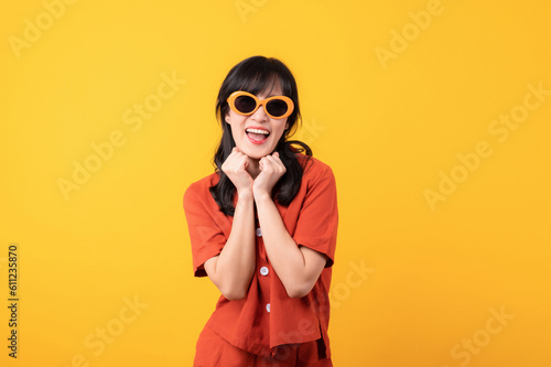 Portrait young beautiful asian woman cute and shy dressed in orange clothes and sunglasses touching cheeks showing shy emotional feeling isolated on yellow studio background.