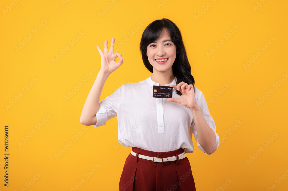 Portrait beautiful young asian woman enterpriser happy smile wearing white shirt and red plants showing okay hand gesture and holding credit card isolation on yellow background. Portrait beautiful young asian woman enterpriser happy smile wearing white shirt and red plants showing okay hand gesture and holding credit card isolation on yellow background.