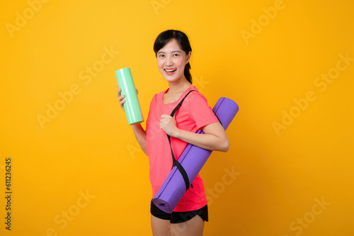 Portrait young asian sports fitness woman happy smile wearing pink sportswear and yoga mat doing exercise training workout against yellow studio background. Healthy wellness lifestyle concept.