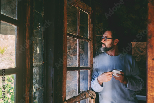 Fotografia One man standing at home chalet drinking tea and looking outside the window with serious thoughtful expression
