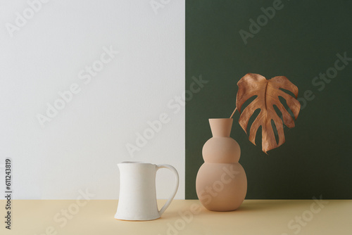Horizontal shot of modern color-blocking still life composition of beige vase with dried plant leaf and creamer against white and dark green wall background, copy space photo