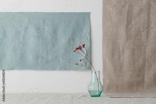 Horizontal shot of minimalistic still life installation with glass vessel and dried red flowers against white wall, gray and blue linen fabric background, copy space photo