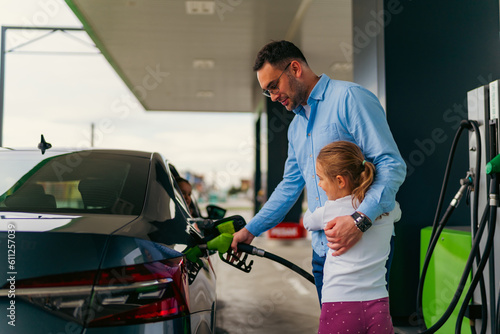 The father pours fuel into the car while his daughter hugs him and keeps him company, younger daughter is sitting in the car