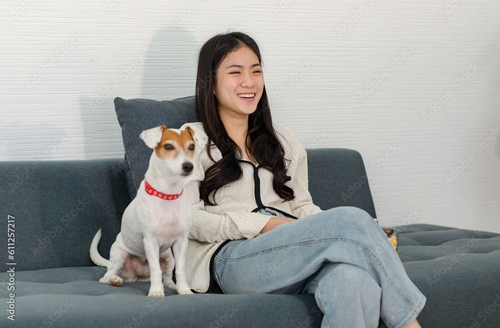 Asian young happy cheerful female owner sitting smiling on cozy sofa couch holding eating popcorn glass bowl snack watching movie with best friend companion dog white short hair jack russell terrier Asian young happy cheerful female owner sitting smiling on cozy sofa couch holding eating popcorn glass bowl snack watching movie with best friend companion dog white short hair jack russell terrier