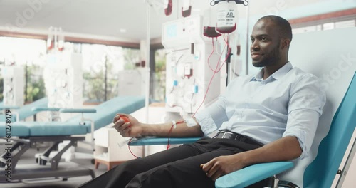 Black Man Donating Blood For People In Need In Bright Hospital. African Male Donor Squeezing Heart-Shaped Red Ball To Pump Blood Through The Tubing Into The Bag. Donation for Children Battling Cancer. photo