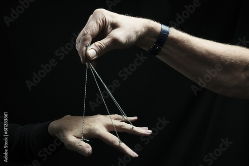 The fingers of a woman's hand are tied with a string. A man's hand holds strings. Dark background.