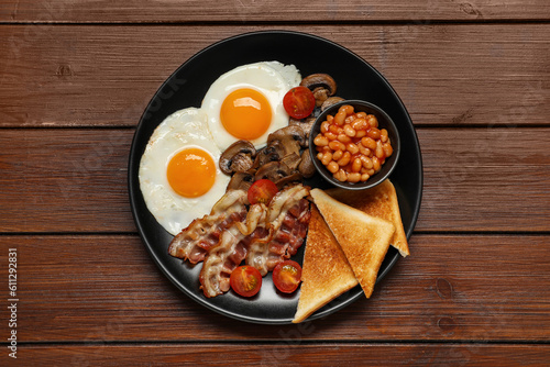 Plate with fried eggs, mushrooms, beans, bacon, tomatoes and toasted bread on wooden table, top view. Traditional English breakfast