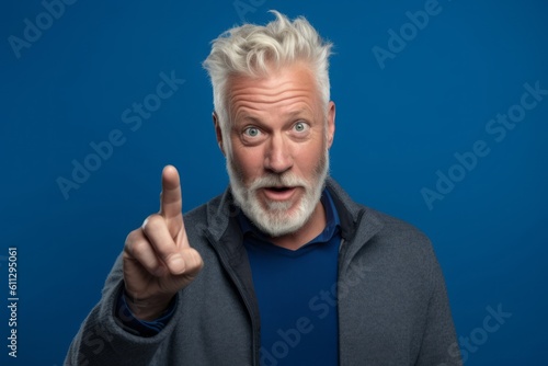 Close-up portrait photography of a beautiful mature man making a i have an idea gesture with a finger up against a royal blue background. With generative AI technology