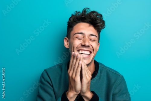 Medium shot portrait photography of a happy boy in his 30s placing the hand over the mouth in a laughter gesture against a turquoise blue background. With generative AI technology