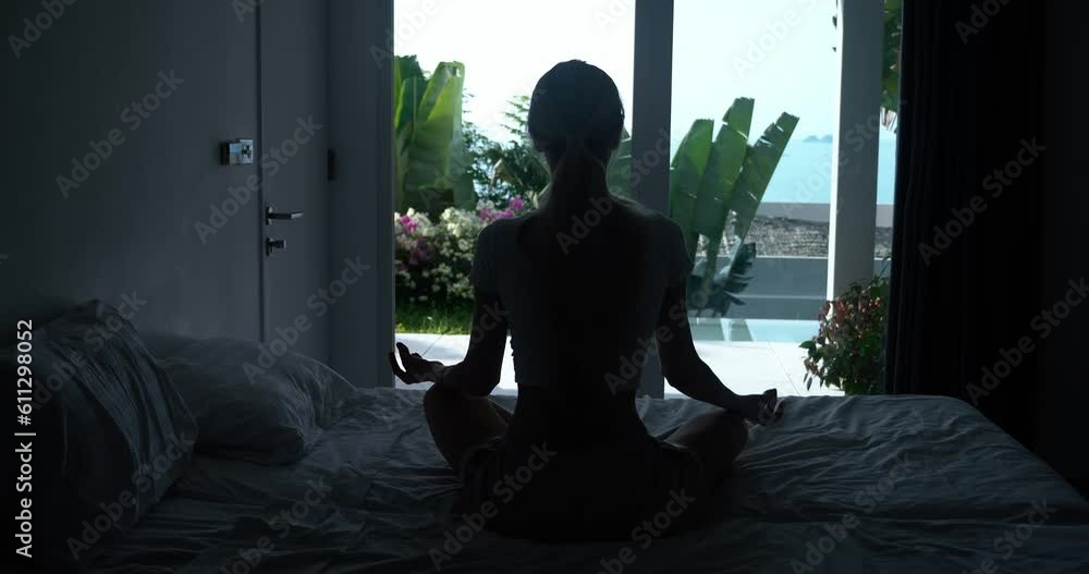 Silhouette woman meditating sitting on bed in bedroom on window background. Calm woman doing yoga exercise at home for stress relief. Female starts day with meditation yoga, motivation for good mood Silhouette woman meditating sitting on bed in bedroom on window background. Calm woman doing yoga exercise at home for stress relief. Female starts day with meditation yoga, motivation for good mood