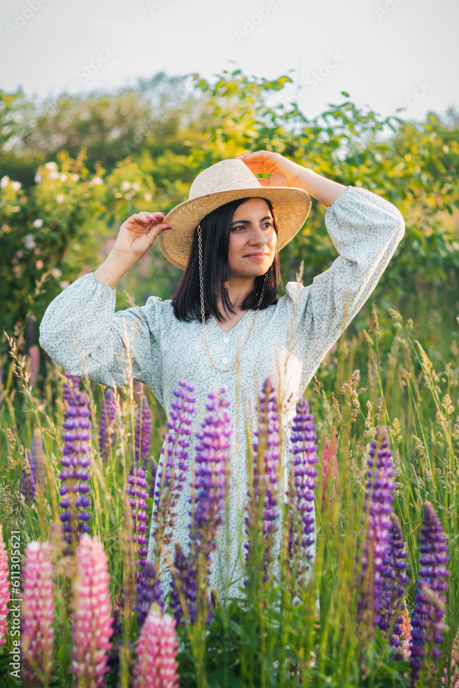 Portrait of young attractive woman in dress and straw hat on the summer meadow with blooming lupin flowers at sunset