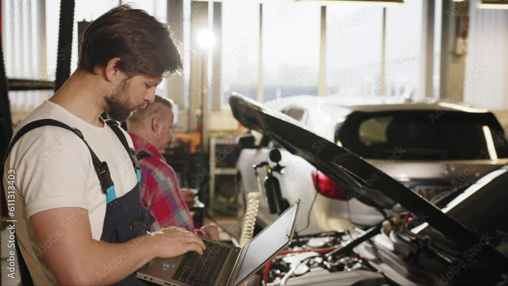 A man in a uniform of a car electrician standing next to car with a diagnostic laptop in auto service workshop garage. Vehicle wiring inspection. Car service, repair, maintenance and people concept A man in a uniform of a car electrician standing next to car with a diagnostic laptop in auto service workshop garage. Vehicle wiring inspection. Car service, repair, maintenance and people concept