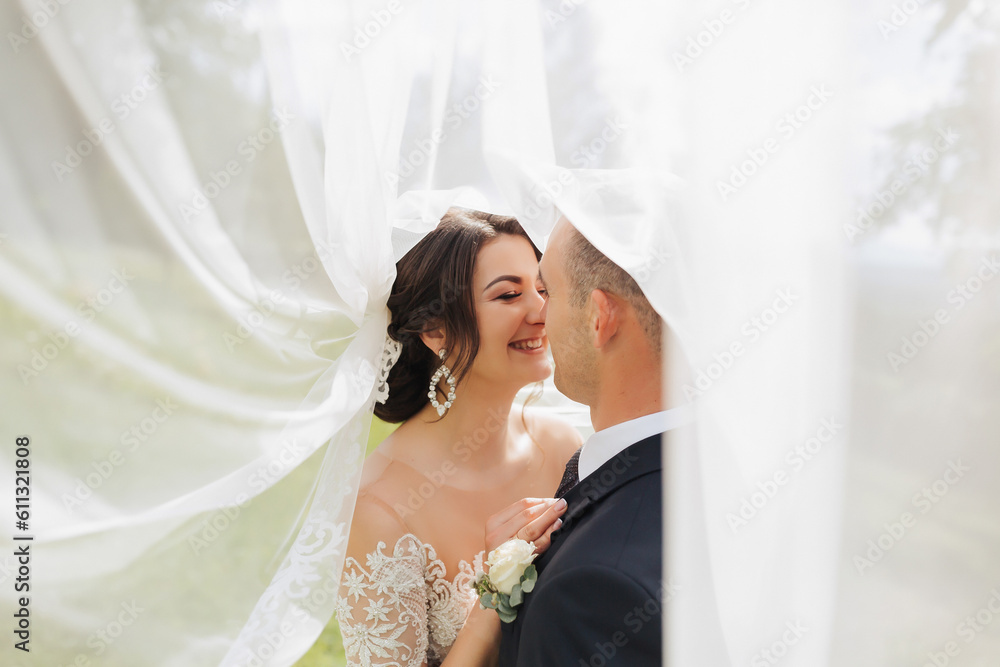 A curly-haired brunette bride in a white dress and a veiled groom embrace and kiss. Portrait of the bride and groom. Beautiful makeup and hair. Wedding in nature A curly-haired brunette bride in a white dress and a veiled groom embrace and kiss. Portrait of the bride and groom. Beautiful makeup and hair. Wedding in nature