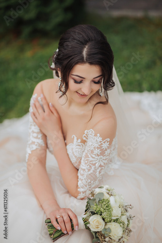 Portrait of the bride in nature. A brunette bride in a white lace dress sits on the grass, poses, holding a bouquet. Beautiful hair and makeup. Open shoulders. Wedding shooting