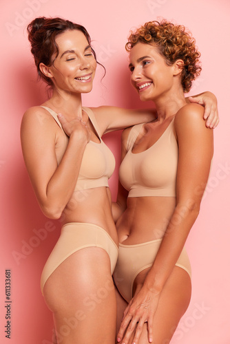 Two pretty women stand against pink background hugging and smiling gently, wearing beige sport bras and panties, relationship concept, copy space