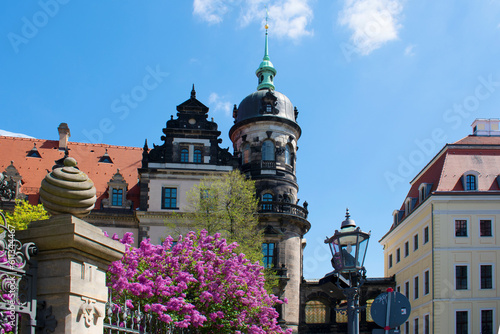 Facade of ancient building with tower, spire and red roof. Old massive fence, street lantern and blooming lilac in the foreground. Sunny day with blue sky. Dresden, Germany, May 2023.