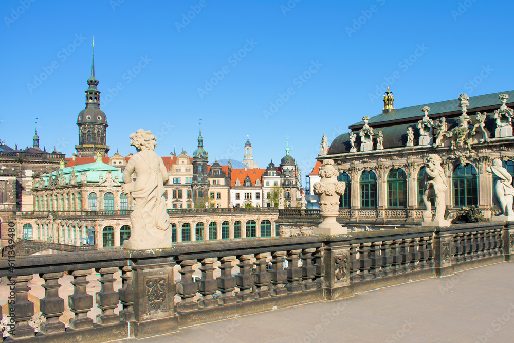 Fototapeta premium View of ancient buildings with towers, spires, red roof, decorative elements and sculptures. Ancient antique fence with statues of boys in the foreground, baroque style. Dresden, Germany, May 2023.