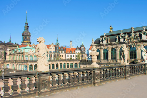 View of ancient buildings with towers, spires, red roof, decorative elements and sculptures. Ancient antique fence with statues of boys in the foreground, baroque style. Dresden, Germany, May 2023.