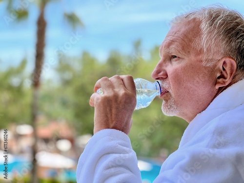 Senior man standing on a resort balcony drinks water from a bottle 