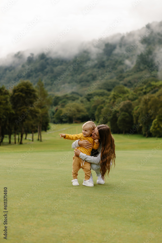 Obraz premium Happy young woman hugs and cuddles her baby while sitting on a green lawn with a beautiful view of the nature garden. Family outdoor recreation, happy family