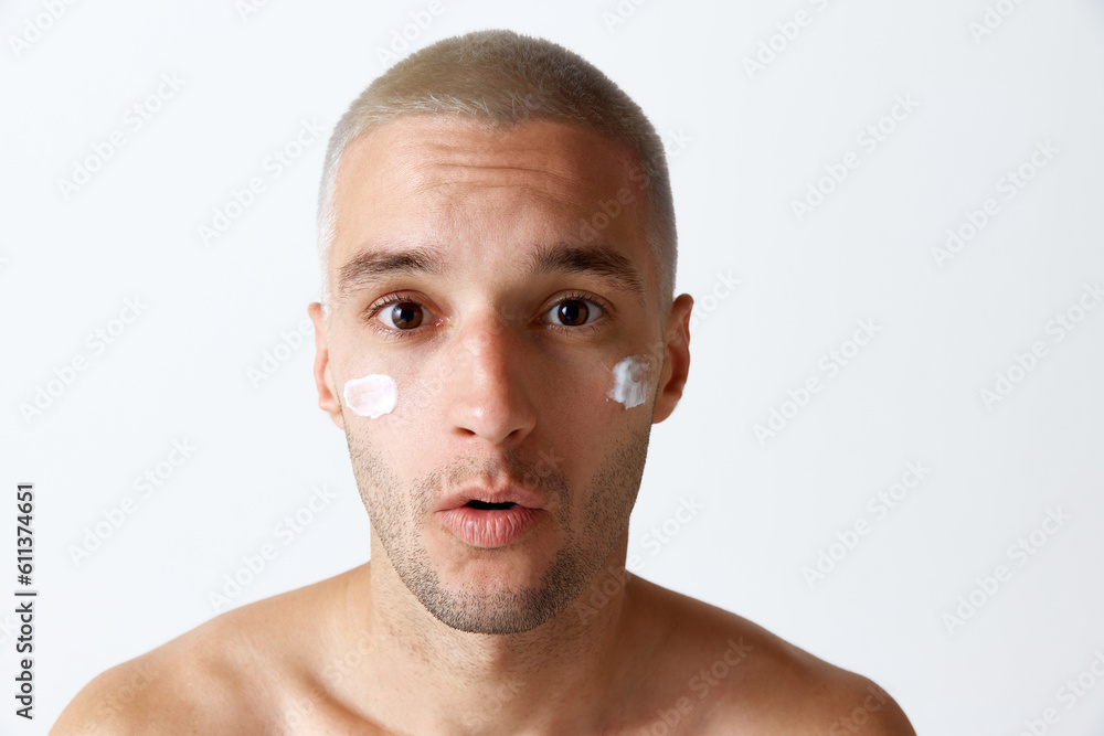 Obraz premium Portrait of young handsome man taking care after skin with moisturizing cream against white studio background. Concept of men's beauty, skincare, cosmetology, spa, health. Copy space for ad