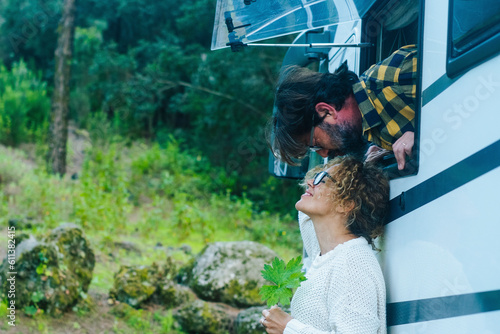 Happy adult cople have fun and love kissing outside a camper van parked in the green nature. Free parking in natural scenic place with trees. Alternative vacation and sweetness. Man and woman