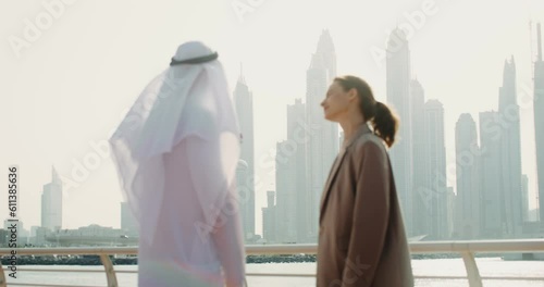 A young man in Arabic traditional white clothes shows his European colleague modern skyscrapers, and then they shake hands photo