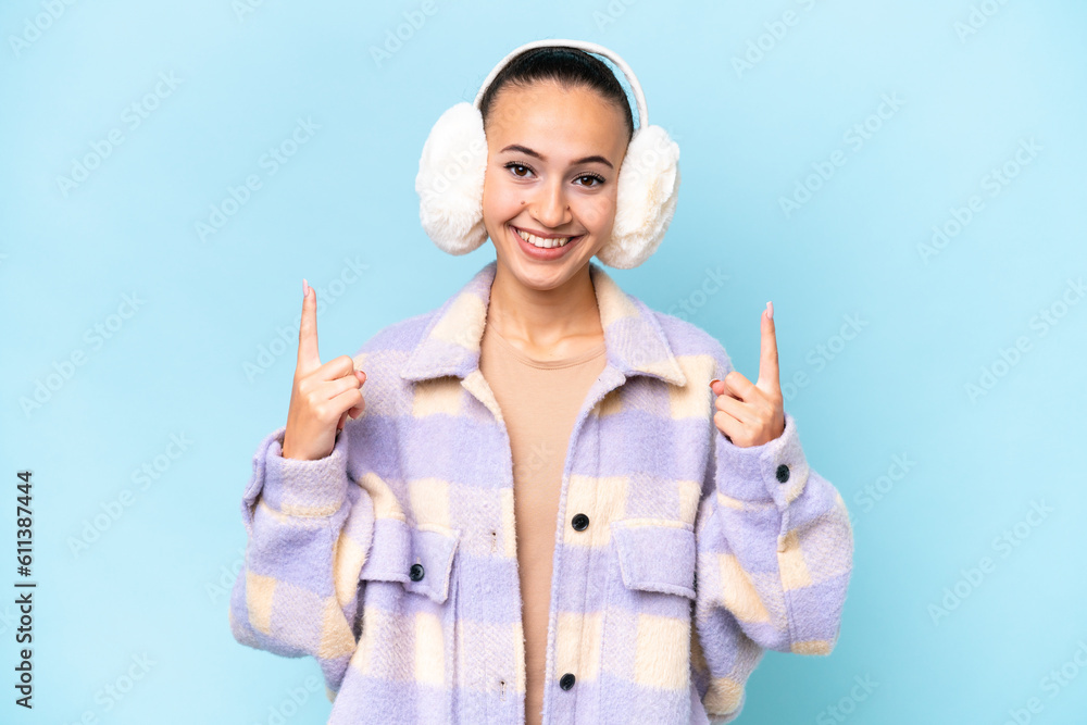 Young Arab woman wearing winter muffs isolated on blue background pointing up a great idea Young Arab woman wearing winter muffs isolated on blue background pointing up a great idea