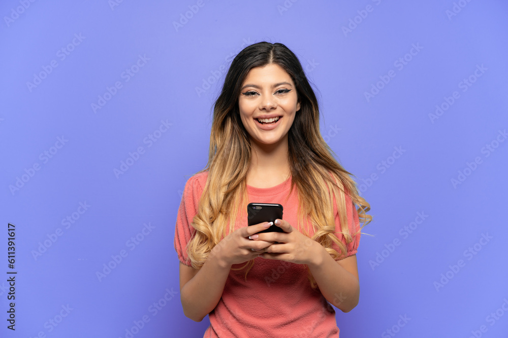 Young Russian girl isolated on blue background looking at the camera and smiling while using the mobile Young Russian girl isolated on blue background looking at the camera and smiling while using the mobile