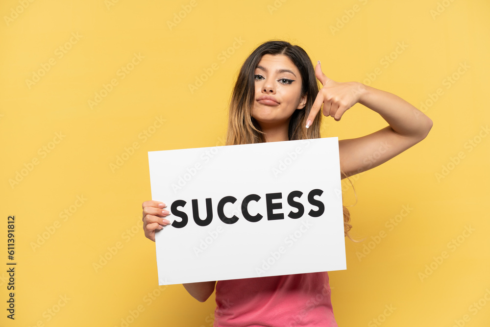 Young Russian girl isolated on yellow background holding a placard with text SUCCESS and pointing it Young Russian girl isolated on yellow background holding a placard with text SUCCESS and pointing it