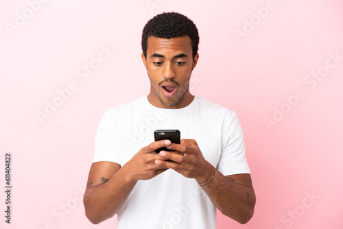 African American handsome man on isolated pink background looking at the camera while using the mobile with surprised expression