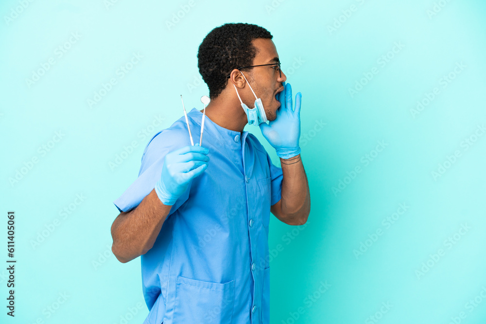 African American dentist holding tools over isolated blue background shouting with mouth wide open to the side African American dentist holding tools over isolated blue background shouting with mouth wide open to the side